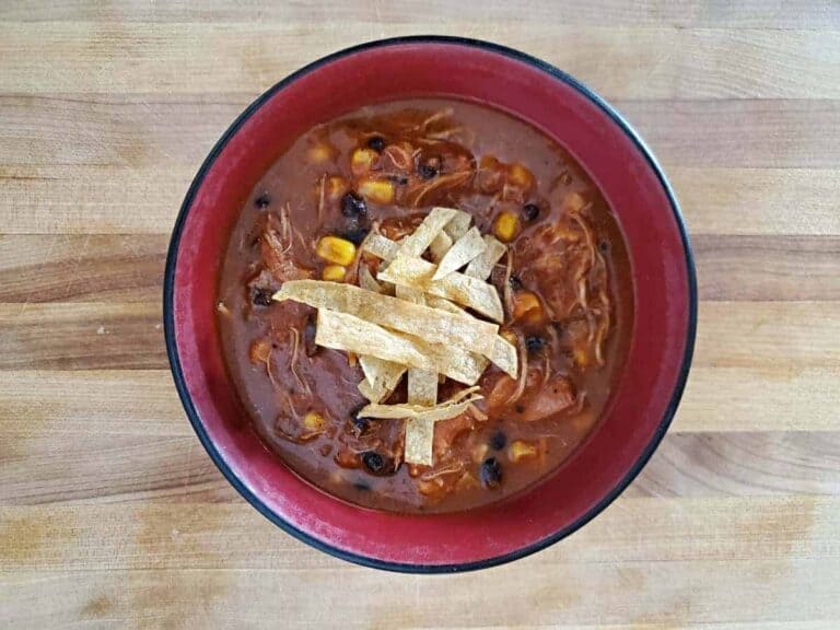 Overhead shot of chicken tortilla soup in a red bowl on wooden table with tortilla strips on the soup.