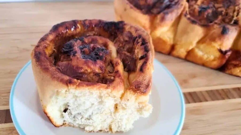 Image shows a three quarters angle shot of a Savory Pulled Pork Cinnamon Roll on a small plate with the rest of the batch behind it.