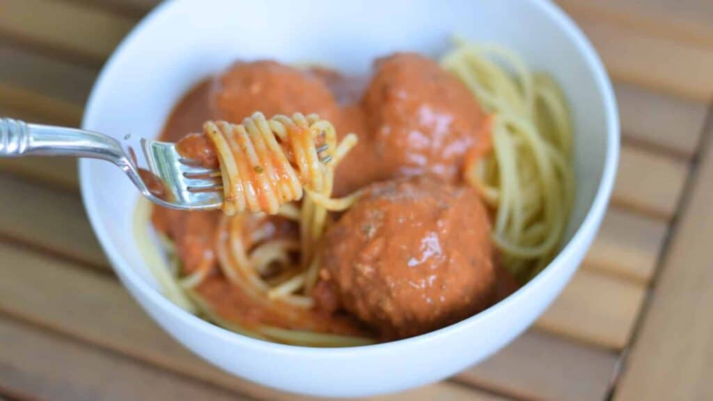 Image shows Spaghetti and meatballs with a fork twirling pasta in a white bowl.