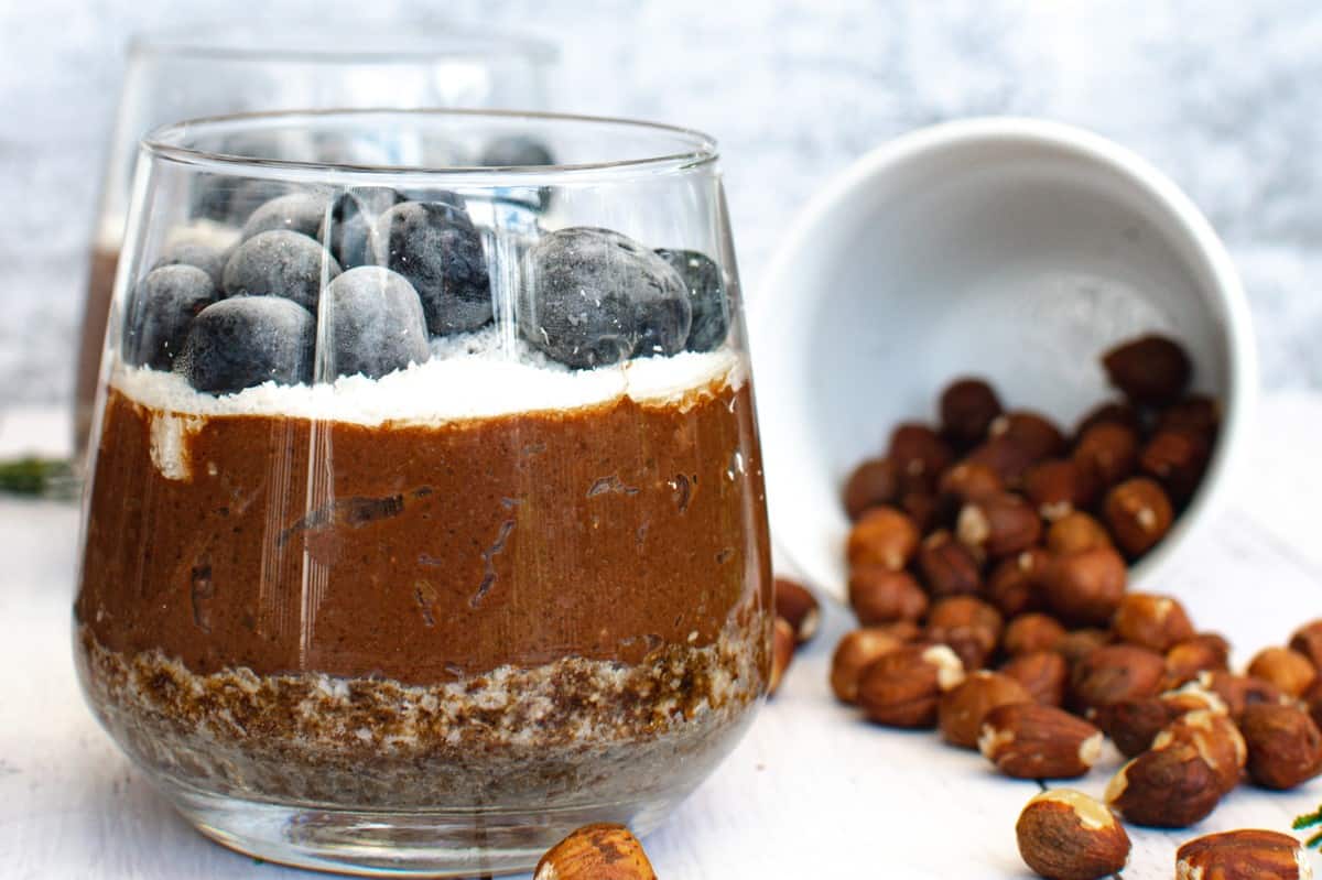 A glass of chocolate pudding with blueberries and hazelnuts.