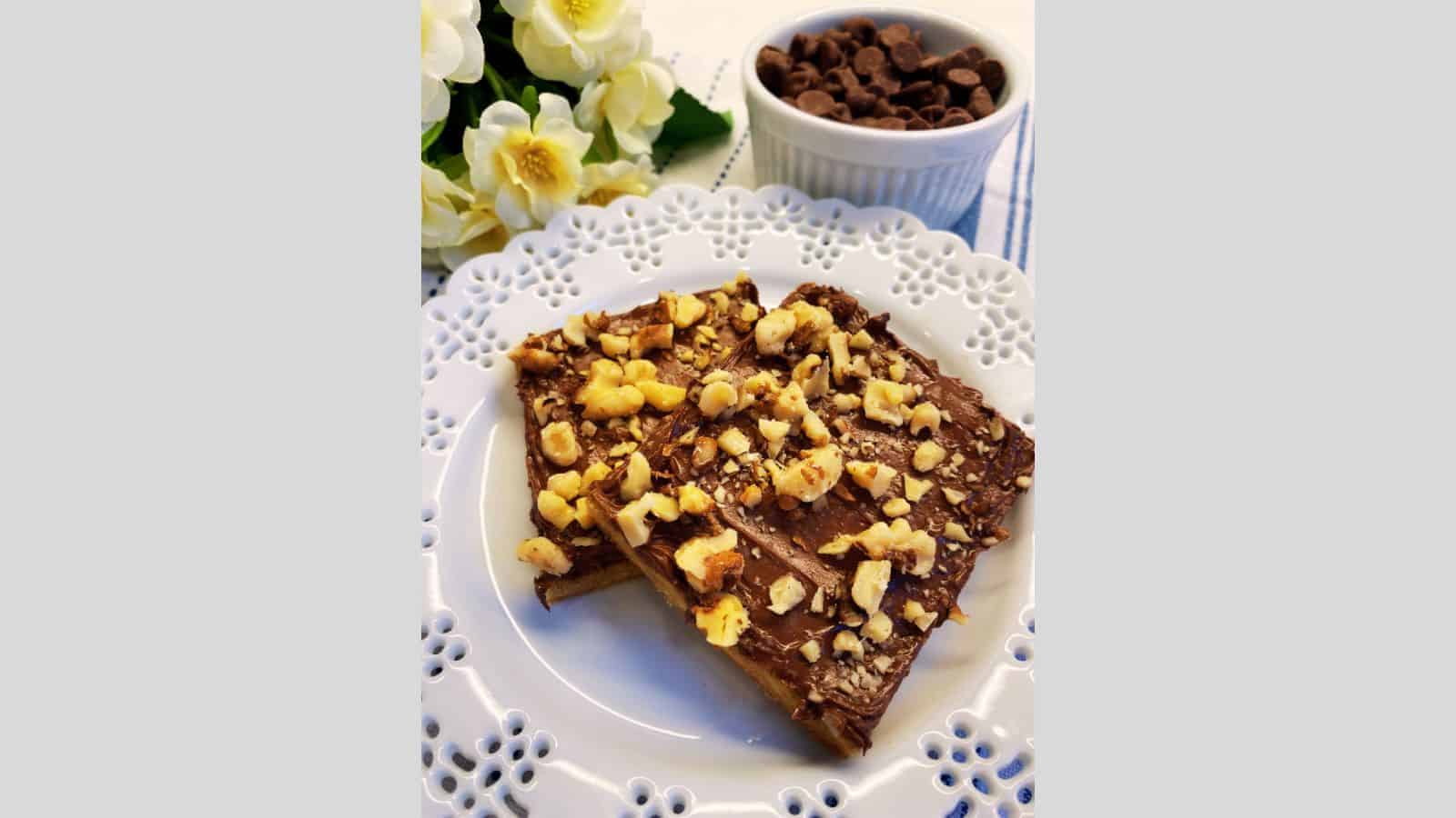 Two toffee cookies on white plate with bowl of chocolate chips in background.