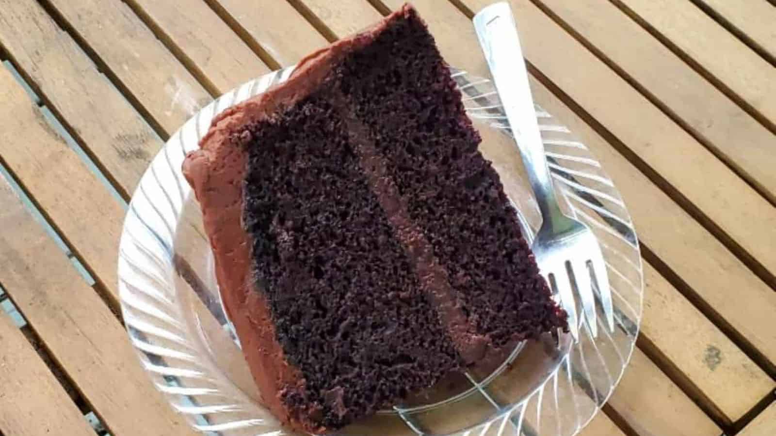Image shows a slice of chocolate frosted chocolate cake on a plate sitting on a wooden table.