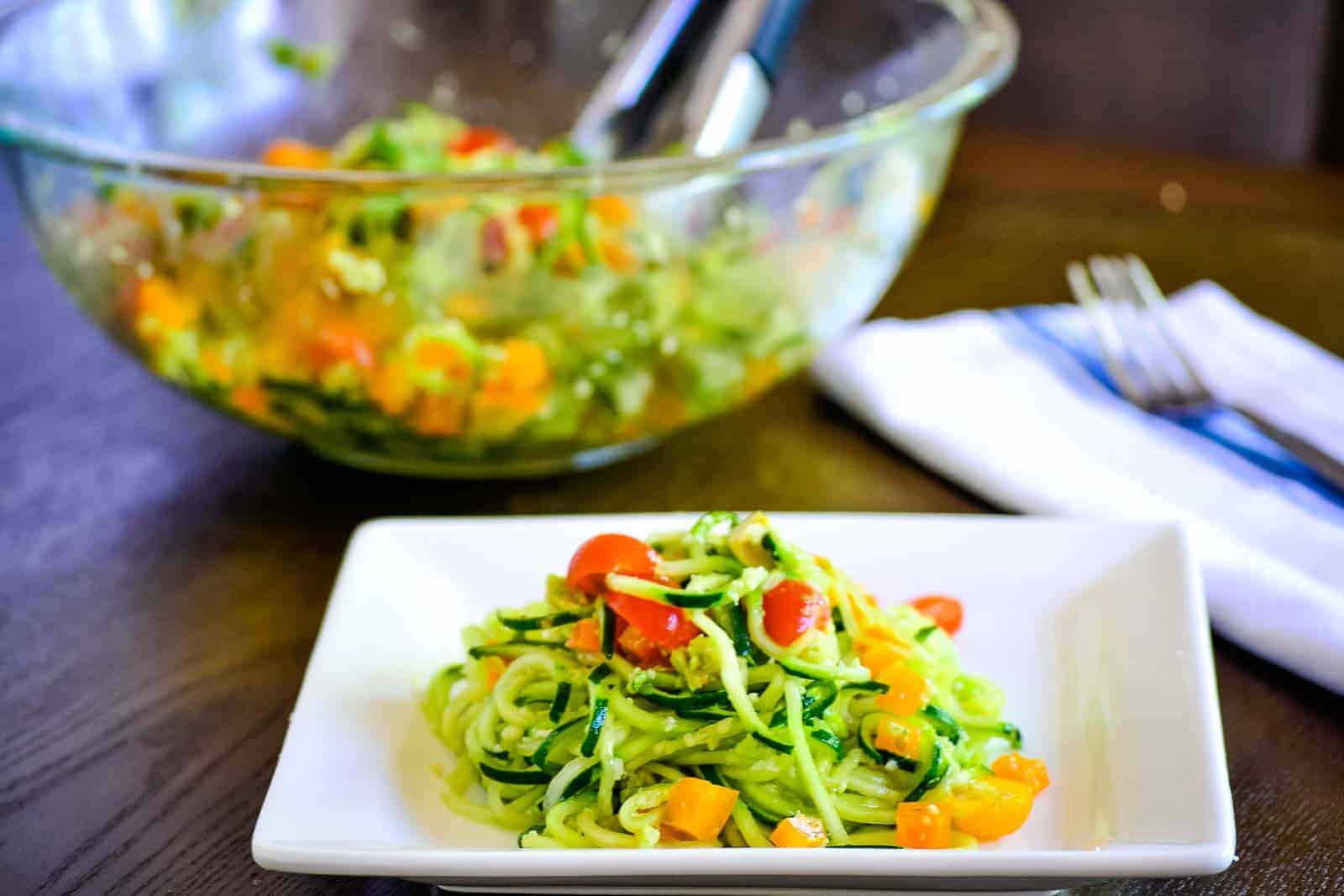 Zucchini noodle salad on a white square plate with a serving bowl in the background.