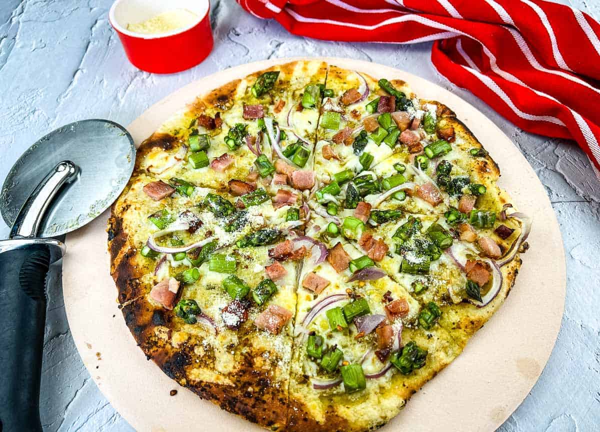 A pizza with ham and asparagus on a pizza stone.