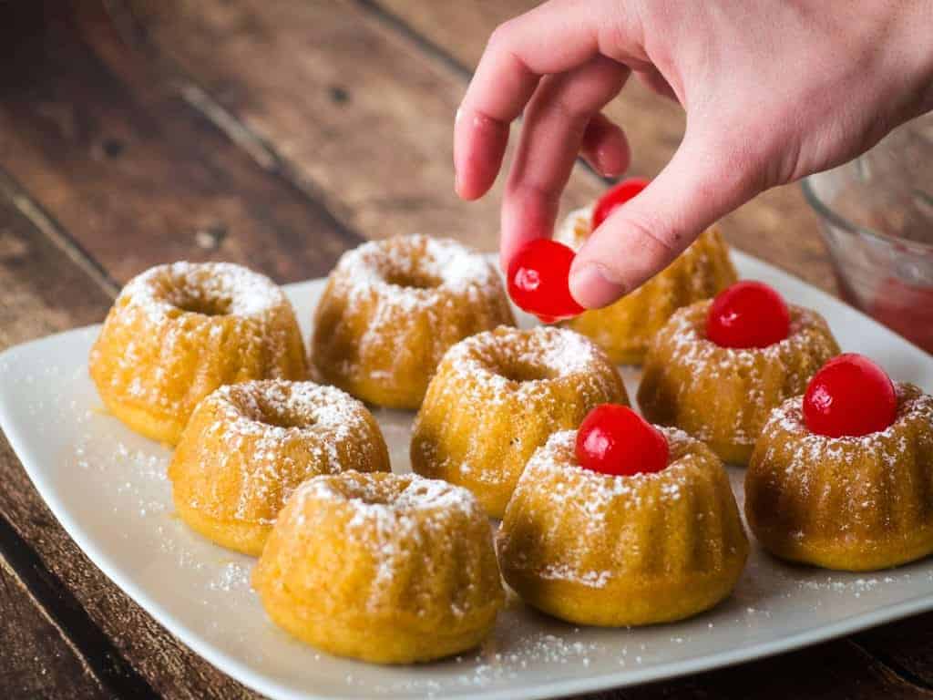 Adding cherries to mini rum cakes on a white plate.