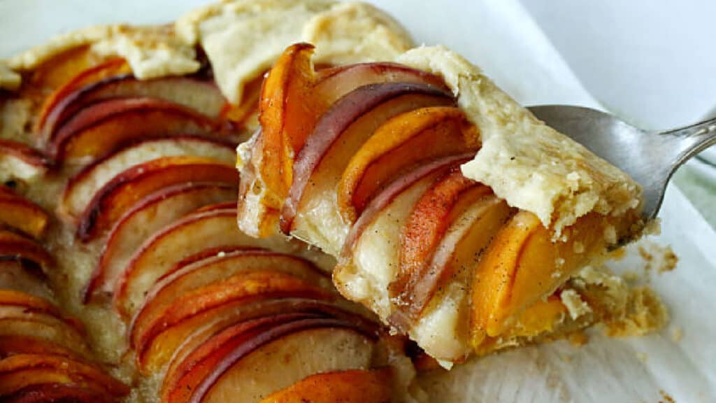 A sweet slice sensation of fruit tart with a golden-brown flaky crust is being lifted by a metal spatula. The tart features thinly sliced peaches and apples arranged in a delicate pattern.