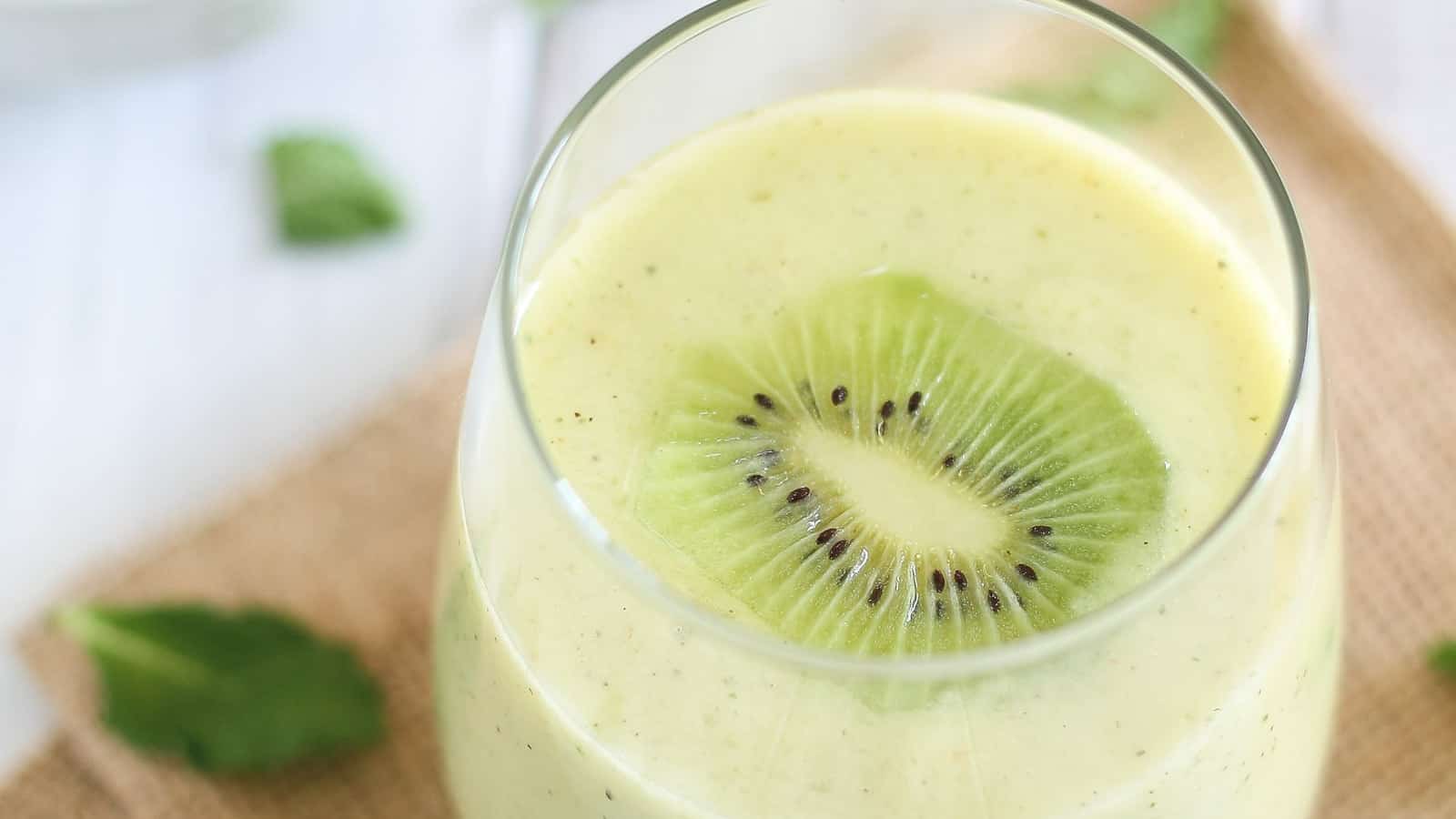 Pineapple kiwi mint smoothie in a glass with kiwi slice garnish.