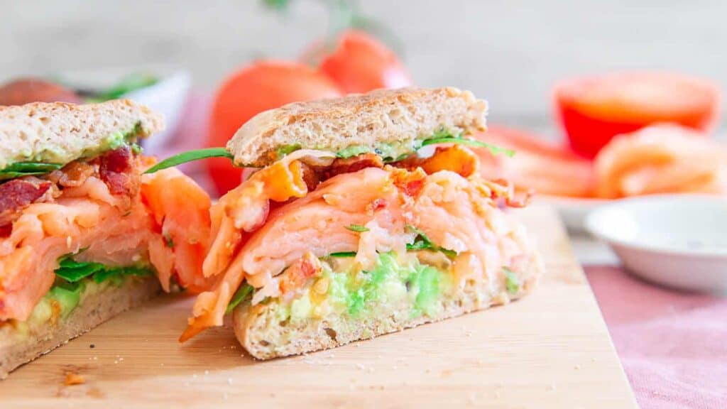 A salmon and avocado sandwich on a cutting board.