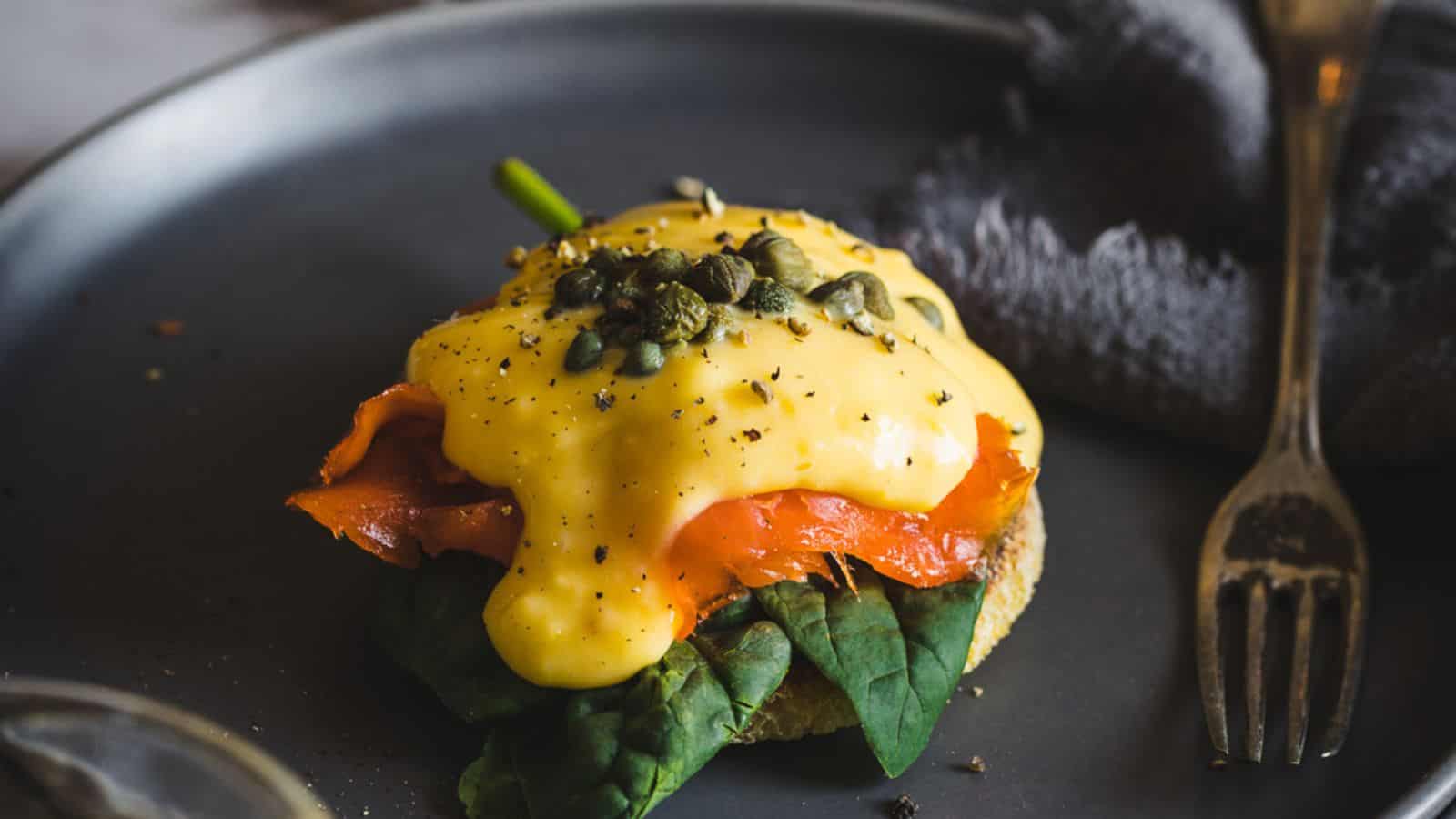 A person holding an eggs benedict with salmon and capers on a plate.