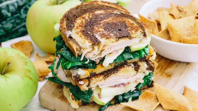 A grilled ham sandwich with greens and apples on a cutting board.