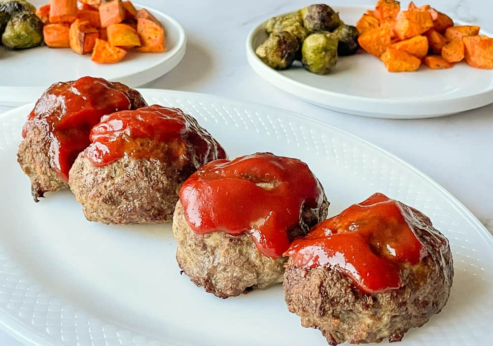 Mini meatloaf on a white platter with roasted brussel sprouts and potatoes in the background.