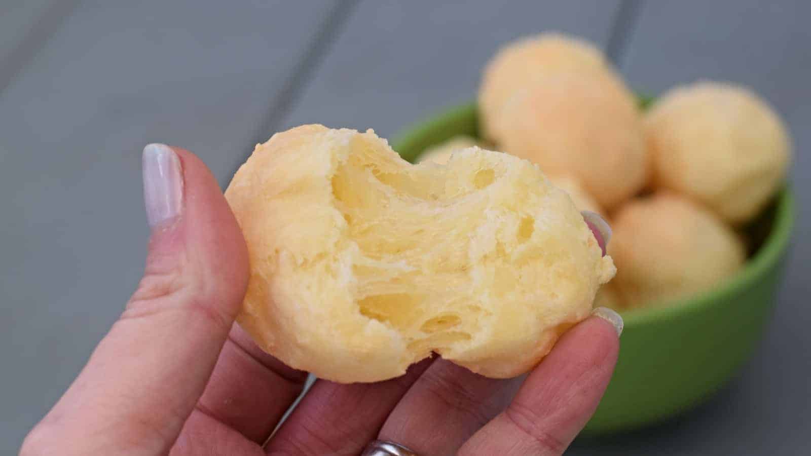 Hand breaking apart brazilian cheese bread with more in a bowl behind it.