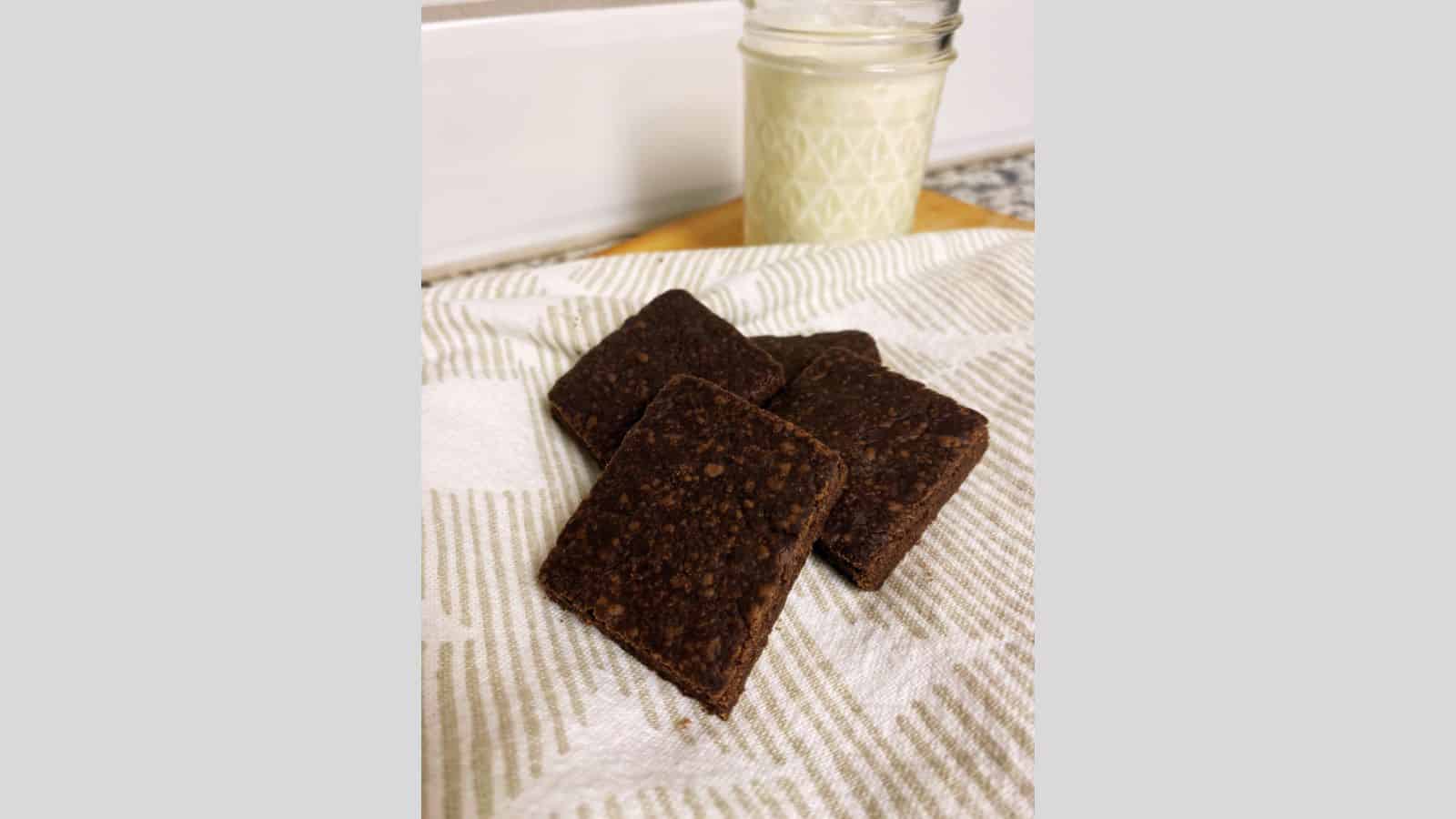 Four chocolate shortbread cookies on white placemat with clear glass of milk in background.