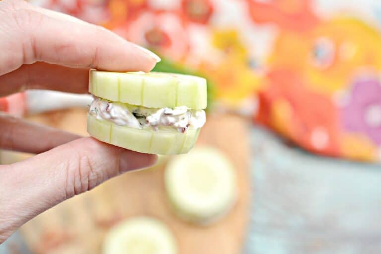 A person holding a cucumber sandwich on a cutting board.