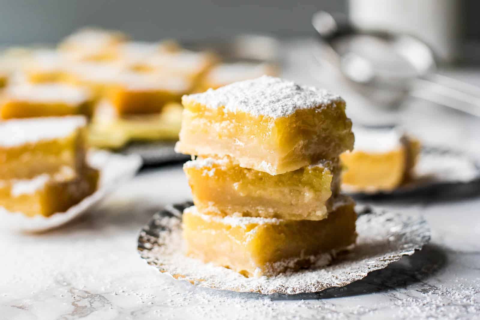 A stack of three lemon bars on a plate with more lemon bars in the background.