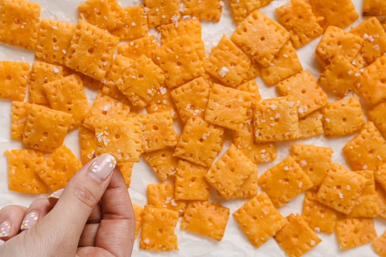 A hand holding a Cheez-It above a baking sheet of more crackers.