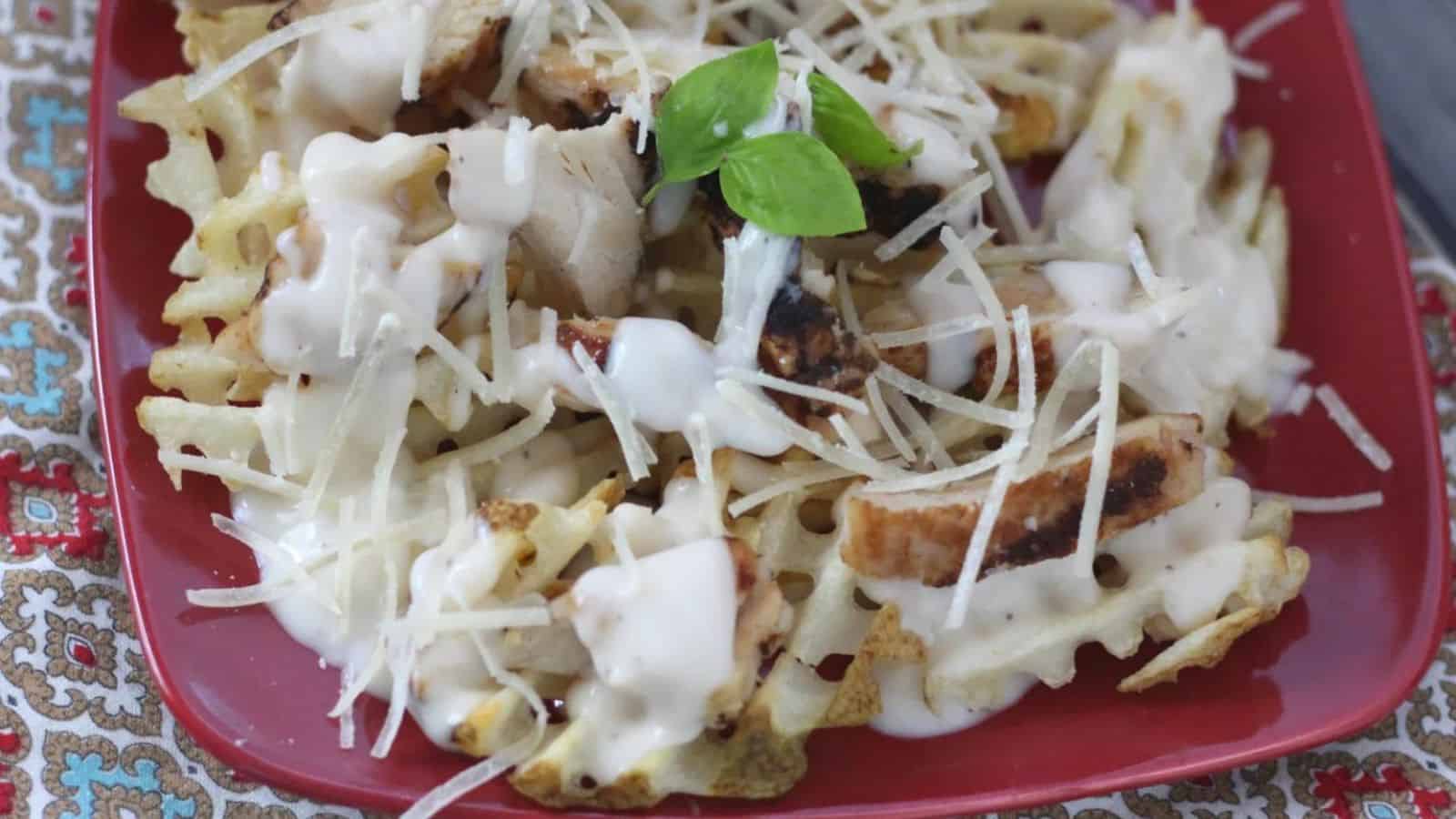 Waffle fries on a red plate topped with grilled chicken, alfredo sauce, and white cheese.