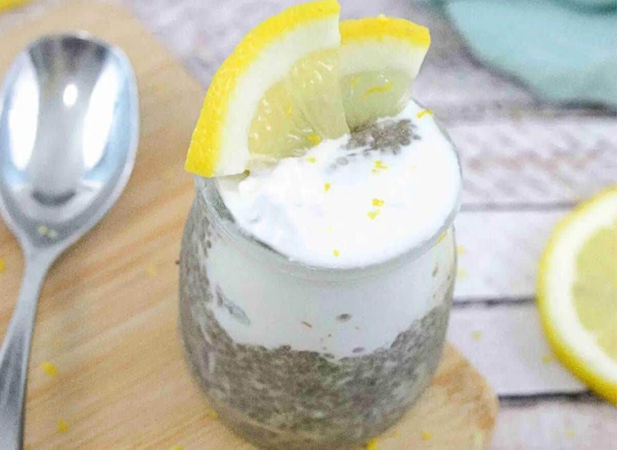 Lemon chia protein pudding topped with coconut cream in a small jar with lemons on top and the jar on a wooden cutting board.