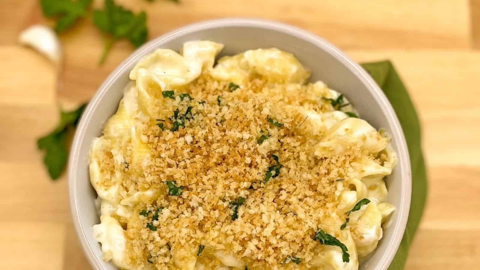 Close up of macaroni and cheese with a breadcrumb and parsley topping in a white bowl with a green linen on a wood cutting board.