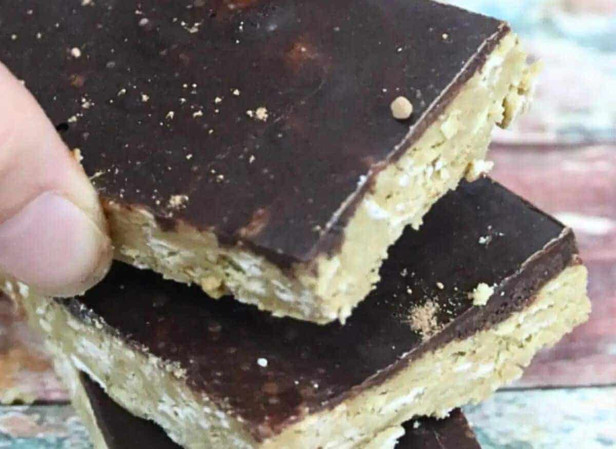 Three nut-free protein bars with chocolate stacked together with a scoop of protein powder and more bars lined up behind it.