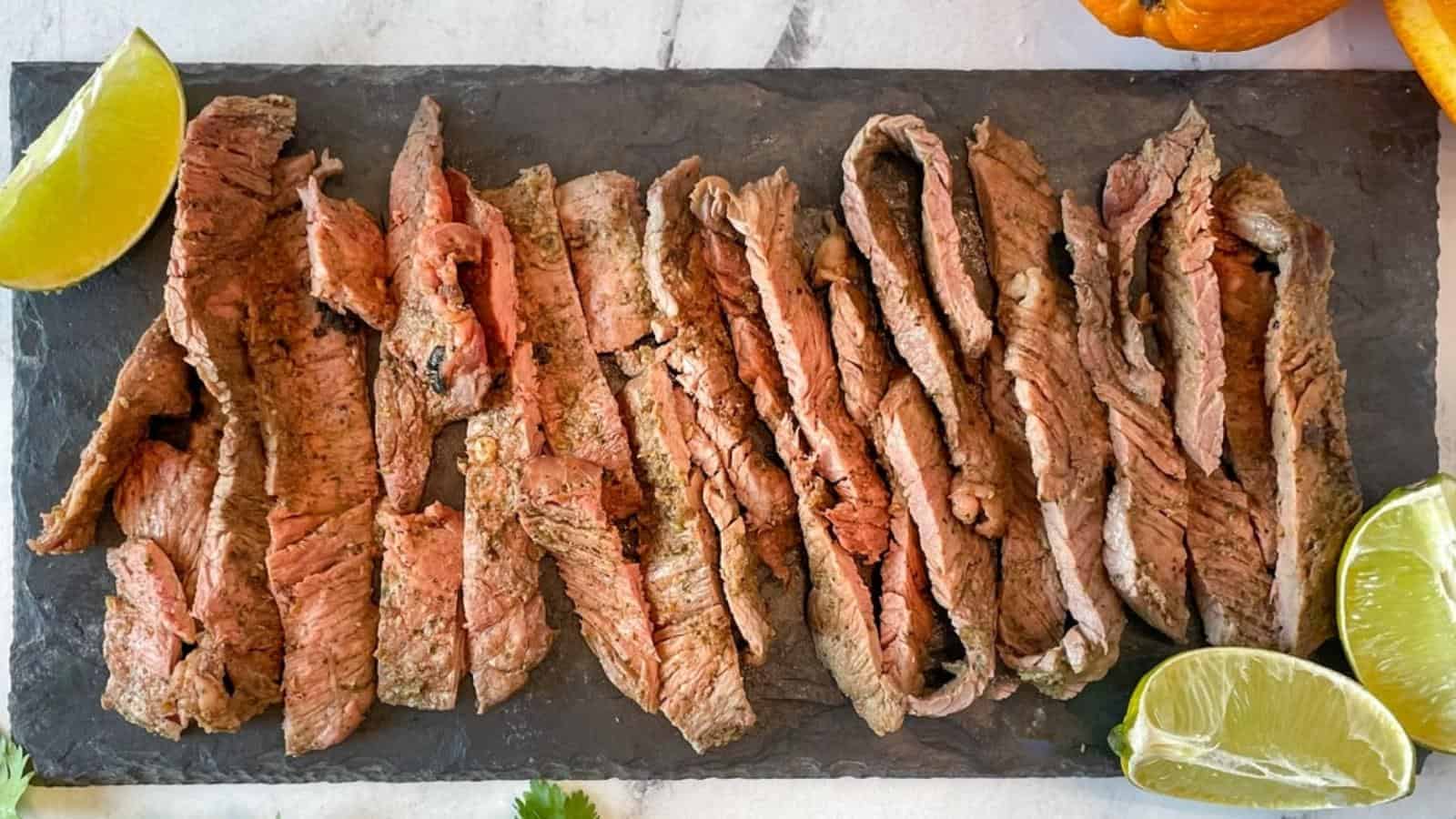 Grilled, sliced ranchera meat is shown on a black stone plate surrounded by lime and orange slices and cilantro.