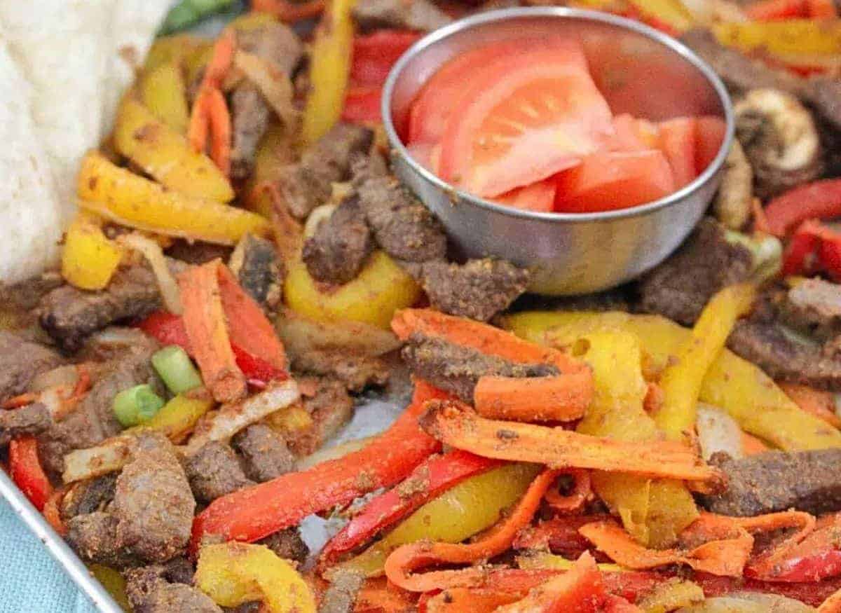 Baking tray with cooked steak, bell peppers, and veggies with a steel bowl of fresh tomatoes in the middle and flour tortillas folded to the side.