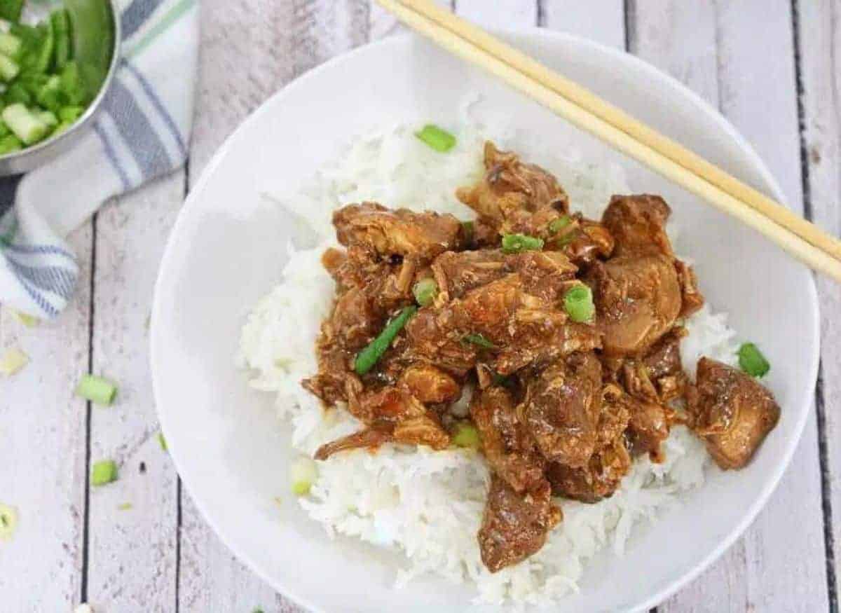 White bowl with rice and teriyaki chicken topped with green onions with chop sticks on the bowl and more green onions behind it.