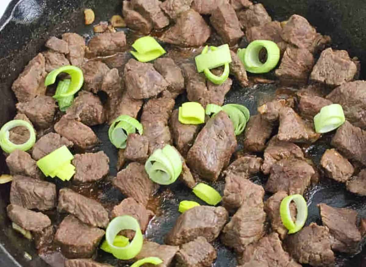 Teriyaki steak bites in a cast iron pan with leeks on top on a blue linen..