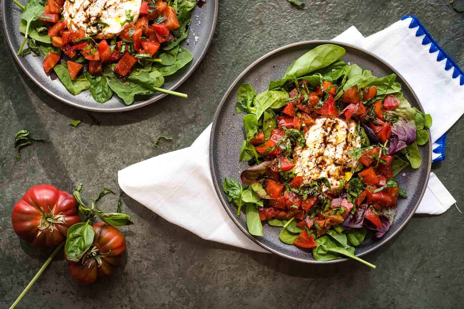 An assembled tomato burrata salad with sliced basil and burrata on a cutting board and a bowl of chopped heirloom tomatoes.