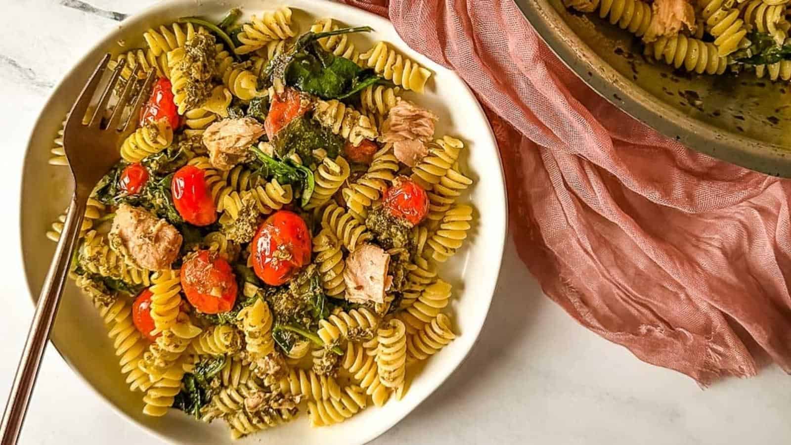 A plate of tuna pesto pasta with cherry tomatoes, a copper fork, and a pink linen next to a pan full of tuna pesto pasta.