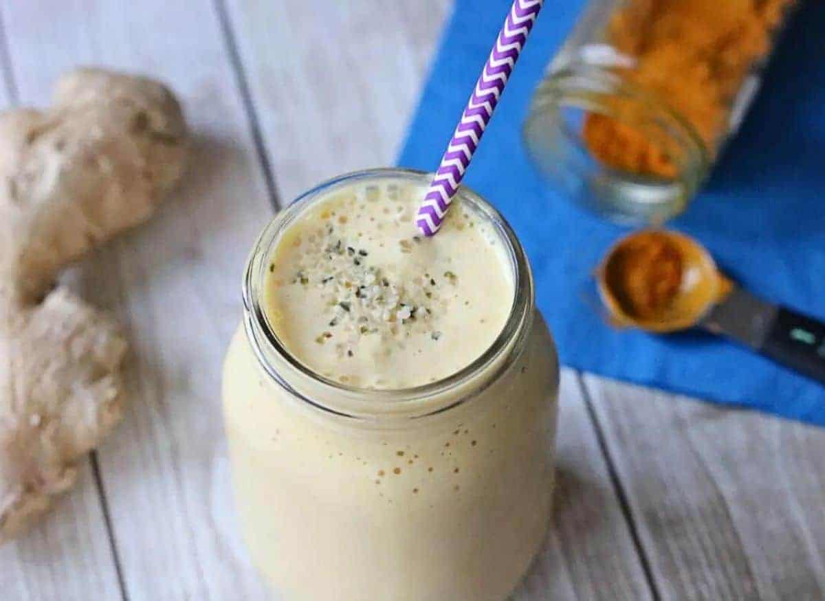 Turmeric peach smoothie in a mason jar with a purple straw. Turmeric root and powder and ginger root around it.