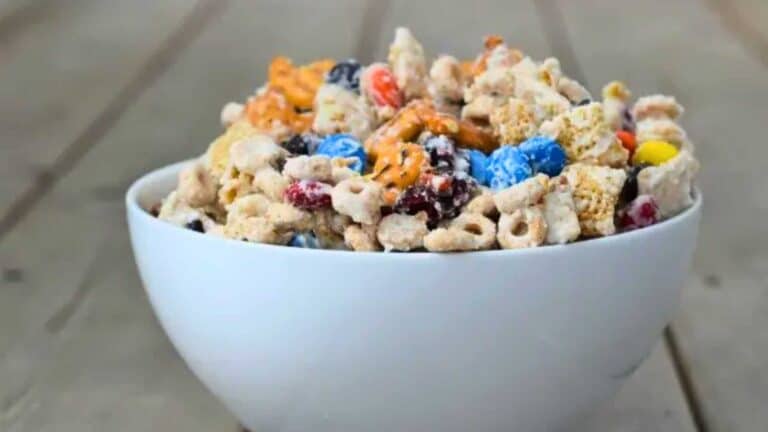 Image shows white chocolate party mix piled into a white bowl on a wooden table.