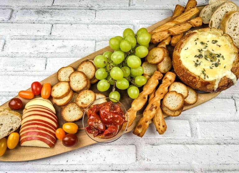 A wooden platter with baked brie in a bowl, cheese, crackers, grapes and bread