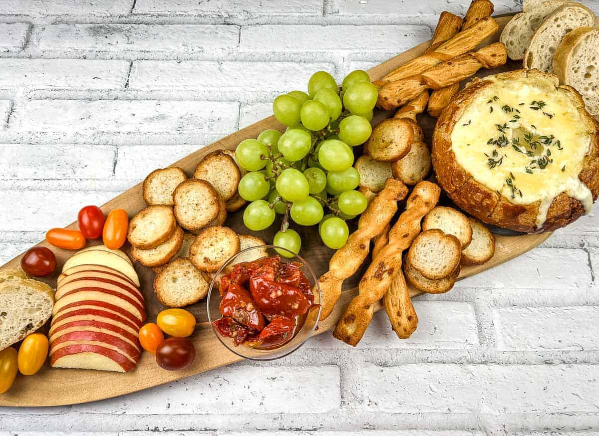 A wooden platter with baked brie in a bowl, cheese, crackers, grapes and bread.