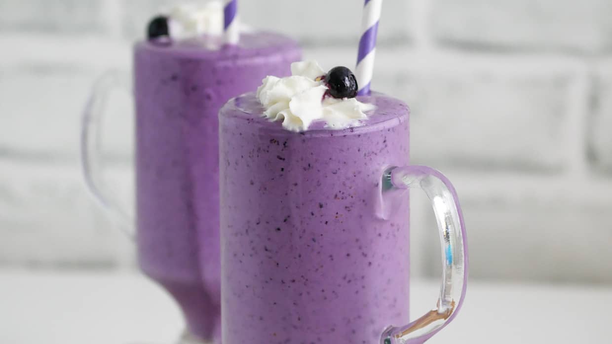 Two glasses of blueberry milkshake with whipped cream and a straw.
