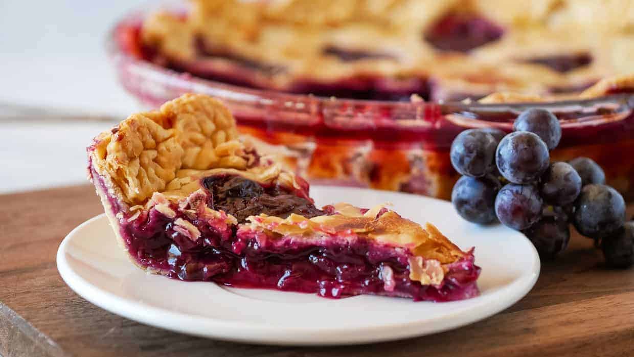 Grape pie on a plate in front of the pie plate with a bunch of grapes.