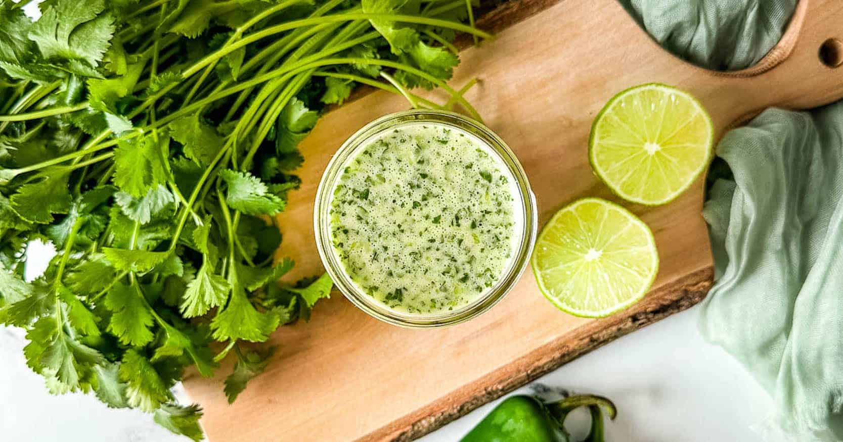 A jar of honey jalapeno lime vinaigrette sits on a cutting board surrounded by limes, cilantro, and jalapenos.