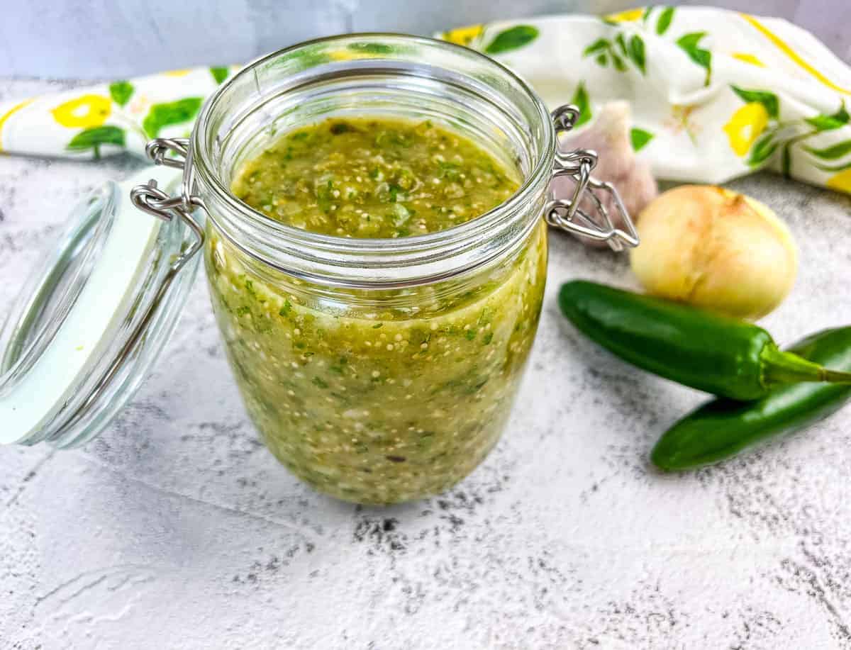 An image of Roasted Tomatillo Sauce in a jar with jalapenos nearby.