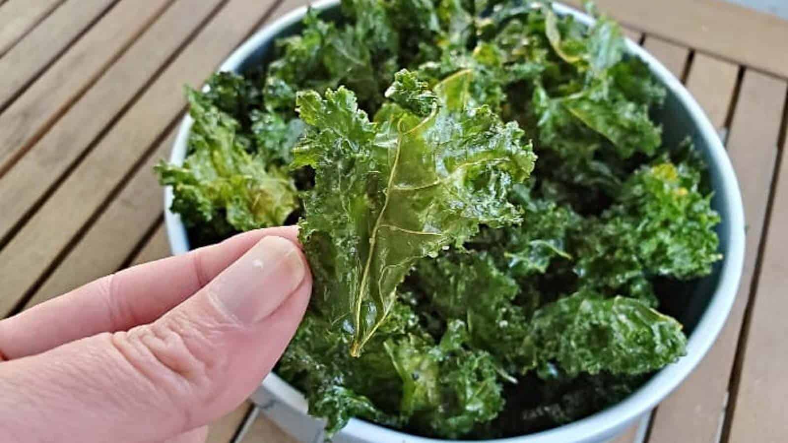 Image shows a hand holding a baked kale chip over a bowl filled with more kale chips.