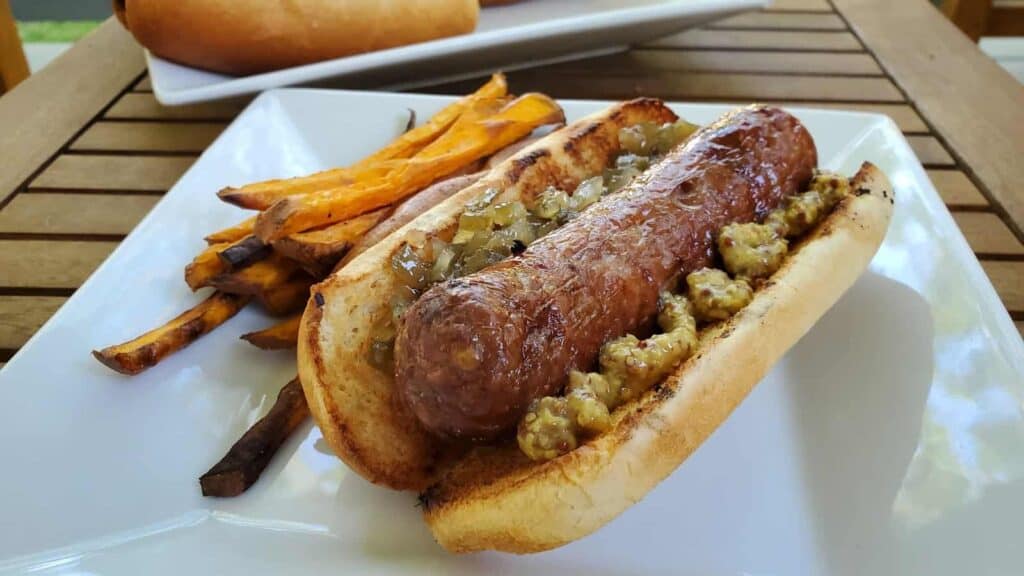 Image shows a beer boiled brat on a bun with sweet potato fries on a plate and more brats in the background.