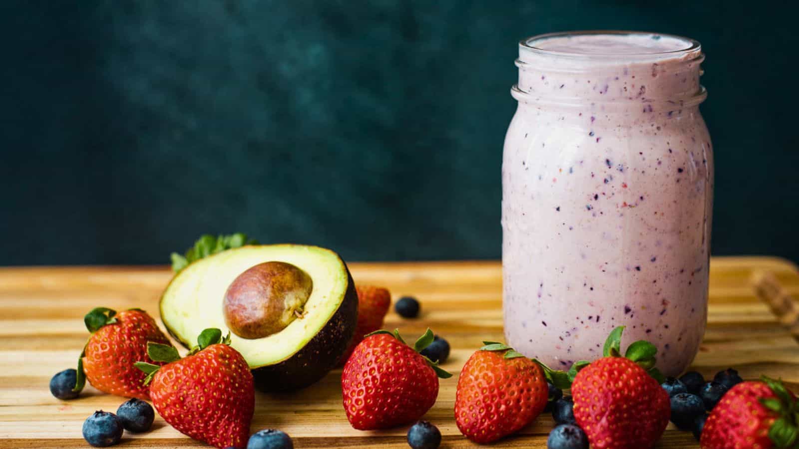 A berry avocado smoothie in a glass mason jar with strawberries, blueberries and avocado on the side.