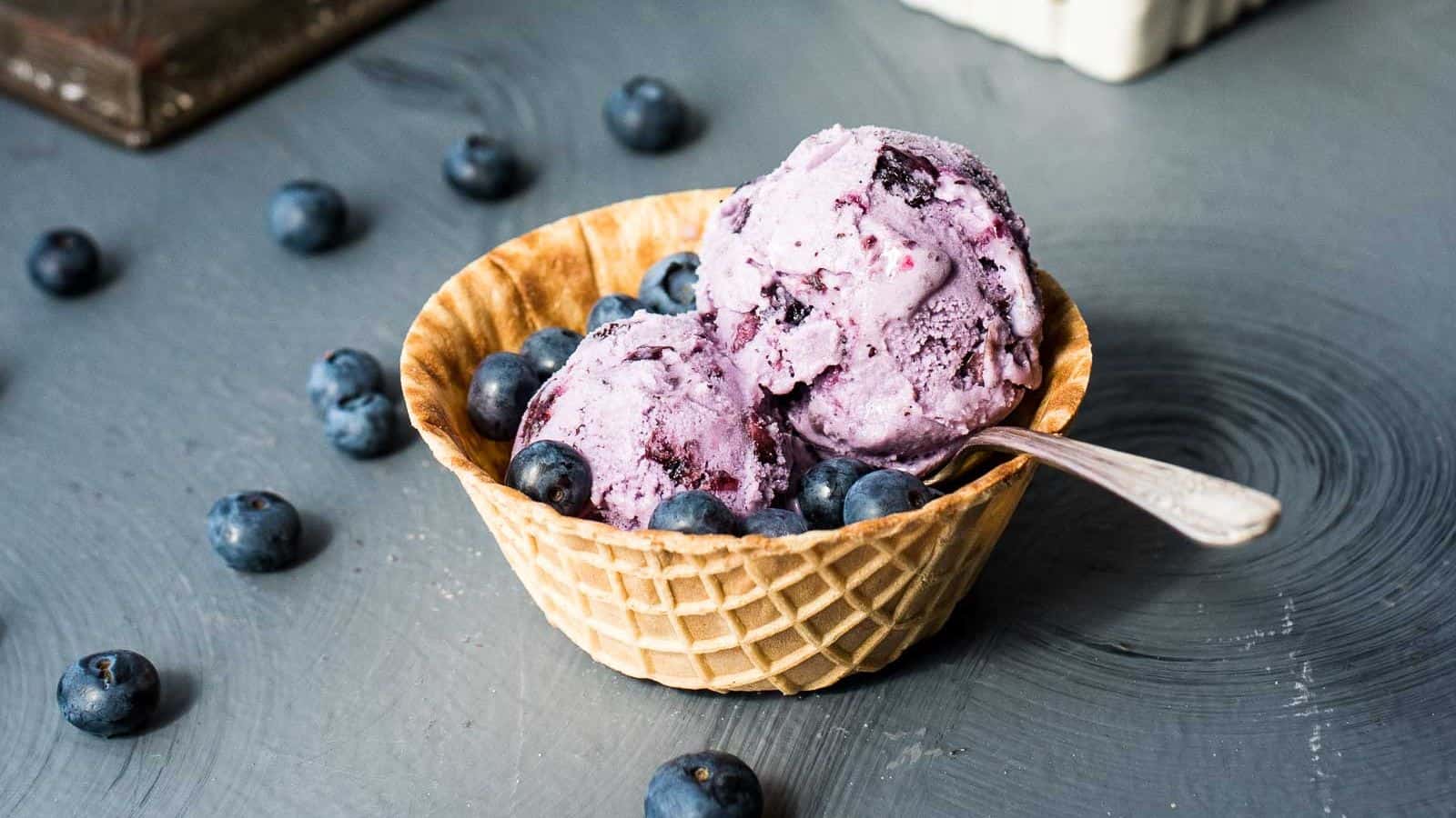 An overhead shot of homemade blueberry ice cream in a metal loaf pan next to waffle bowls, spoons, and a cream colored napkin.