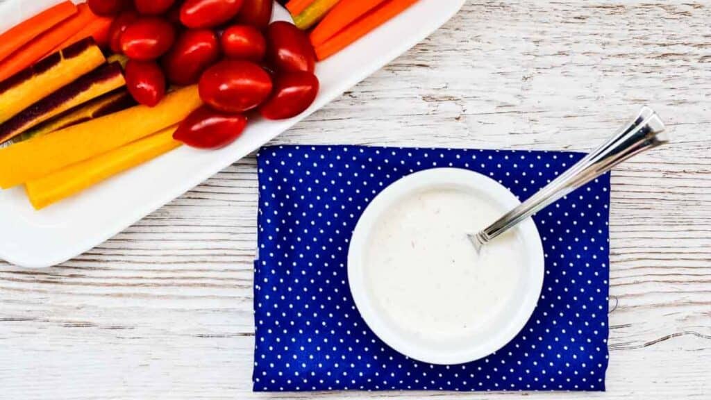 A bowl of creamy garlic dressing next to a plate of carrots, peppers, and cherry tomatoes on a blue cloth.