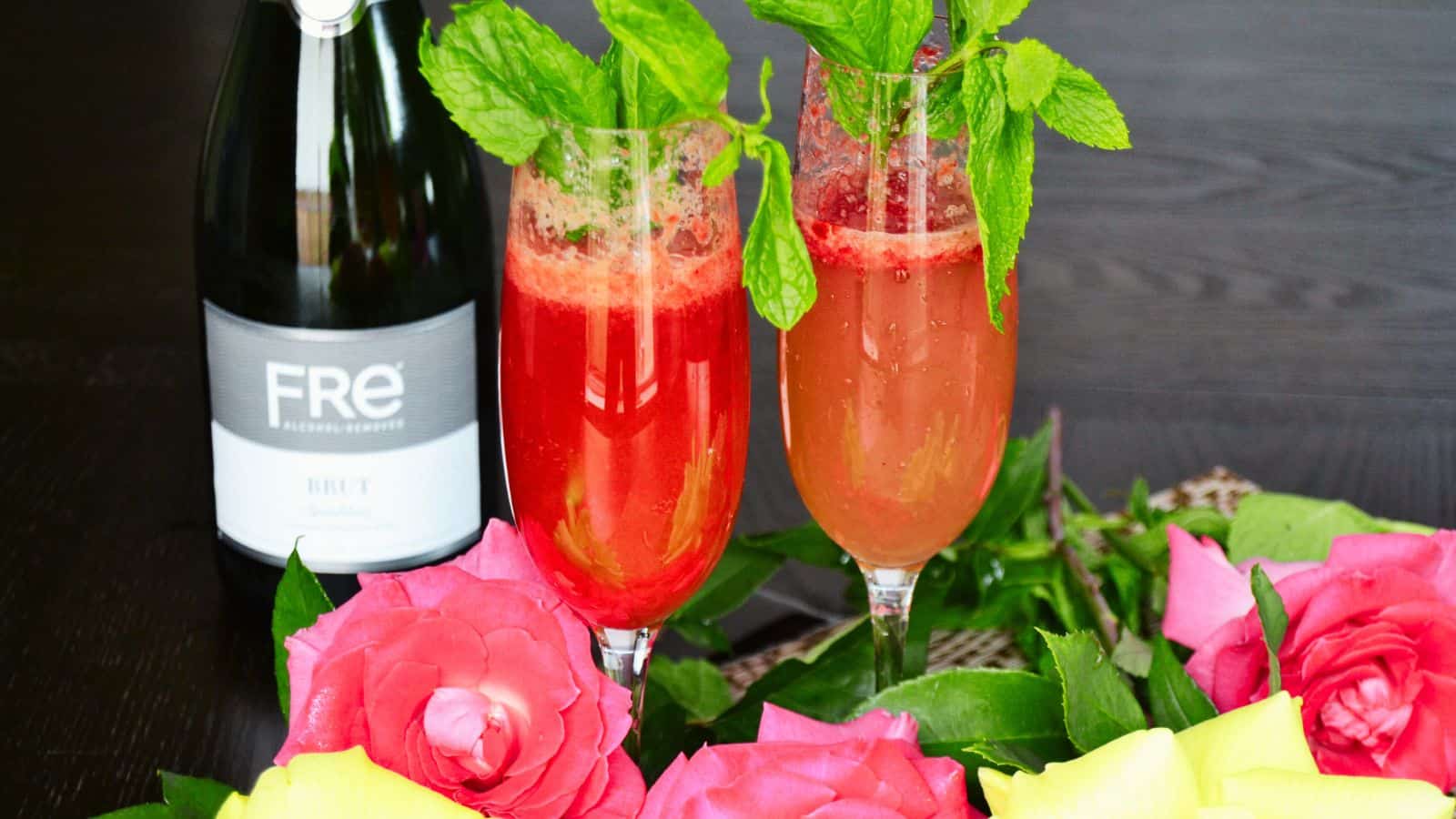 Two raspberry bellini mocktails with roses and a bottle of wine.
