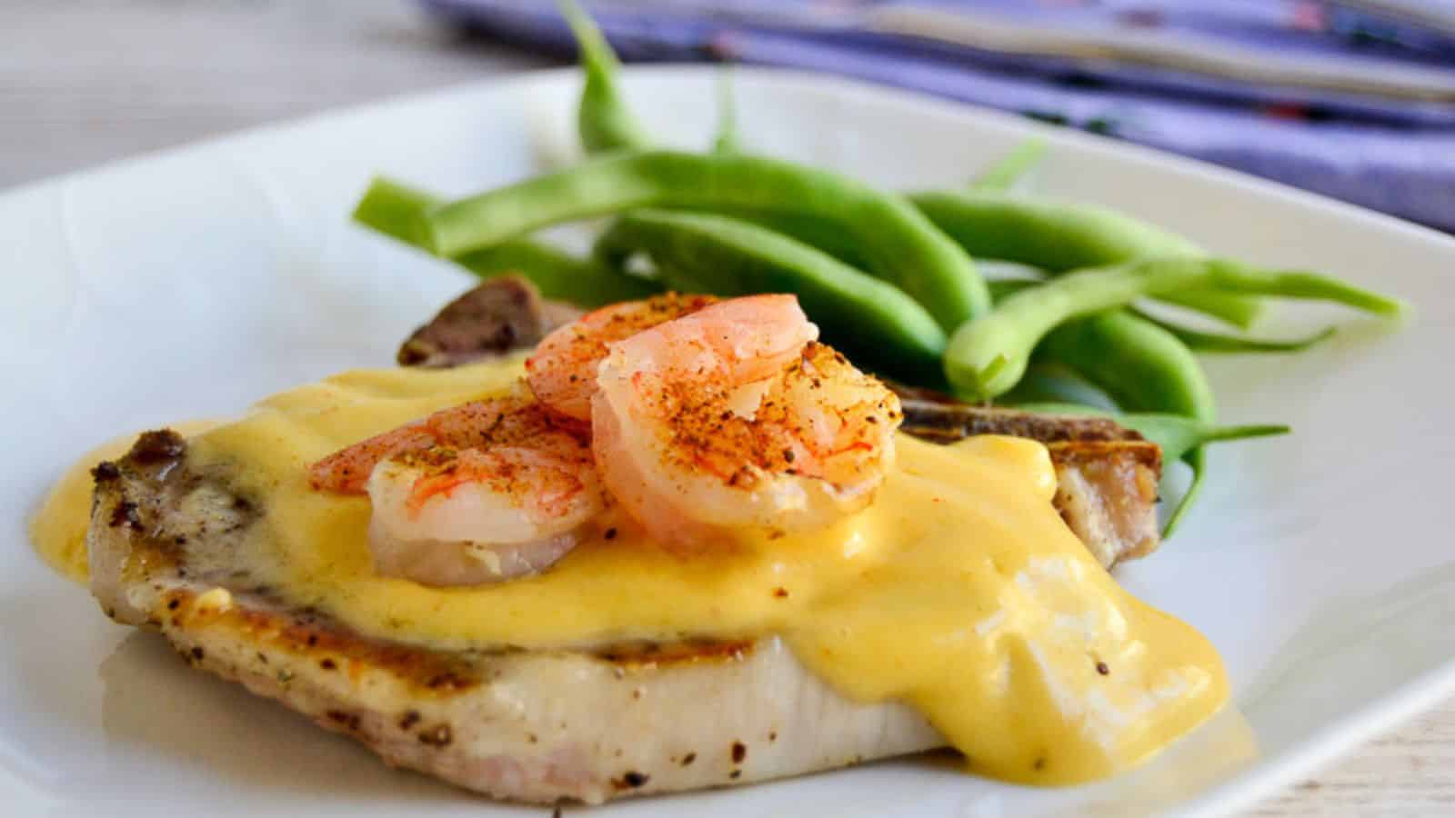 Bone in pork chop topped with hollandaise sauce and shrimp, on a white plate next to green beans.