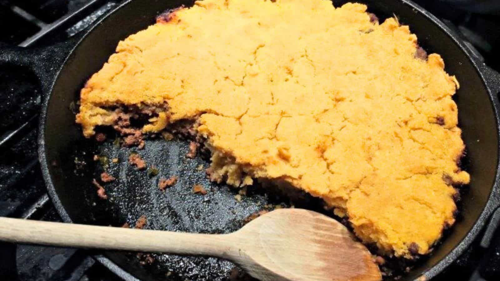 Image shows a partially served ground beef skillet tamales in a skillet with a wooden spoon.