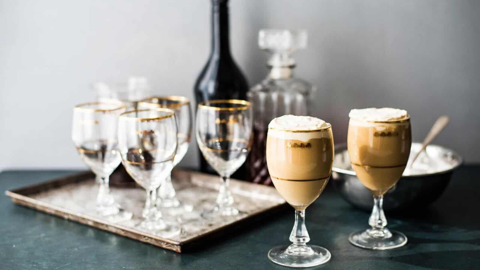 Two glasses of Irish Cream coffee in front of a tray of festive glass and a bar set up for making more.