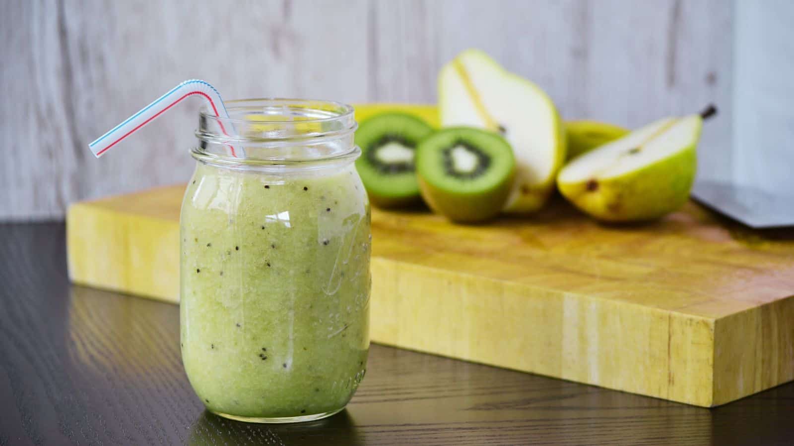A kiwi pear green smoothie in a glass with a kiwi on the rim and a green straw.