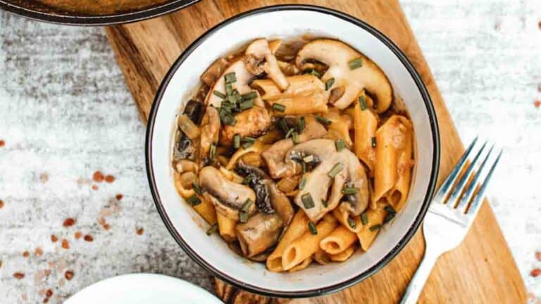 A bowl of pasta with mushrooms and herbs on a wooden cutting board.