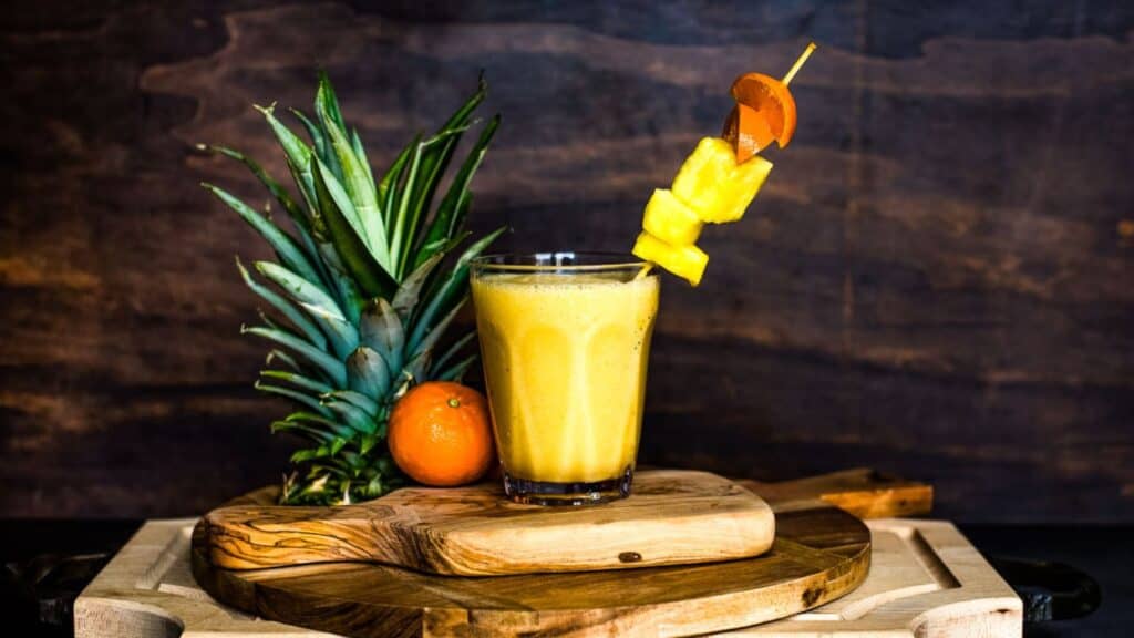 A drink with pineapples and oranges on top of a wooden cutting board.